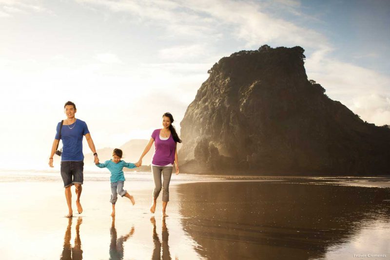 rodzina spacerująca boso w słoneczny dzień brzegiem plaży Piha Beach, w tle skały