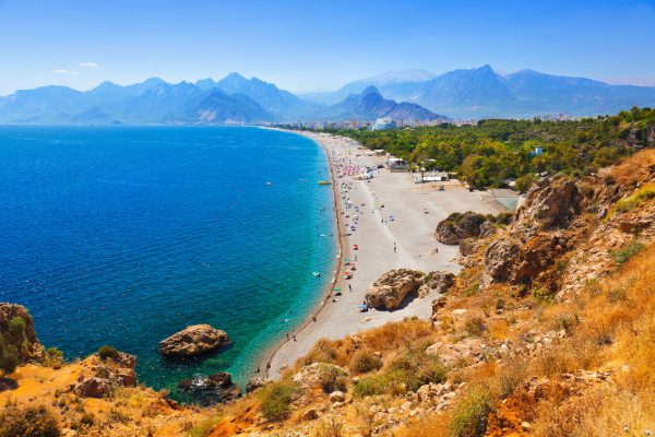 widok wzdłuż wybrzeża w Antalyi, turkusowa woda, szeroka piaszczysta plaża z turystami, w tle wysokie szczyty gór Taurus