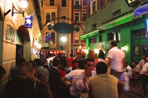 oświetlona, wąska uliczka w Stambule, zatloczona przez siedzących przy barach i restauracjach tubylców