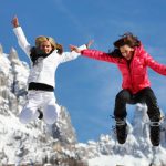 dwie młode kobiety ubrane w zimowe stroje skaczące radośnie na tle ośnieżonych gór we Włoszech