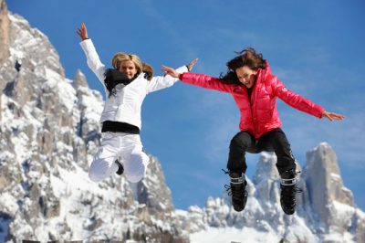 dwie młode kobiety ubrane w zimowe stroje skaczące radośnie na tle ośnieżonych gór we Włoszech